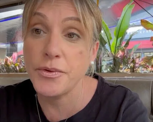 Alison Nightingale, a woman with light hair and pearl earrings, sits indoors near a window with plants outside. She is wearing a black top and looking slightly to the side.